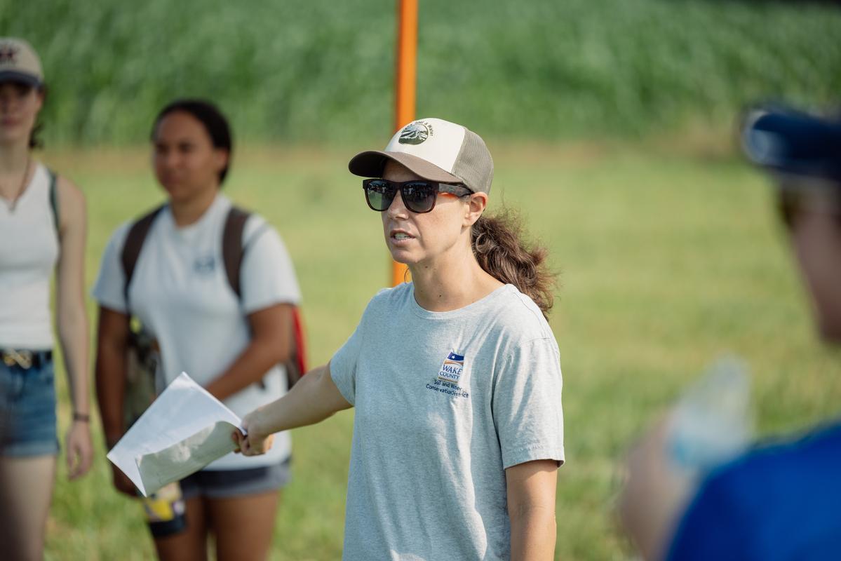 Woman standing in field holding papers talks to students