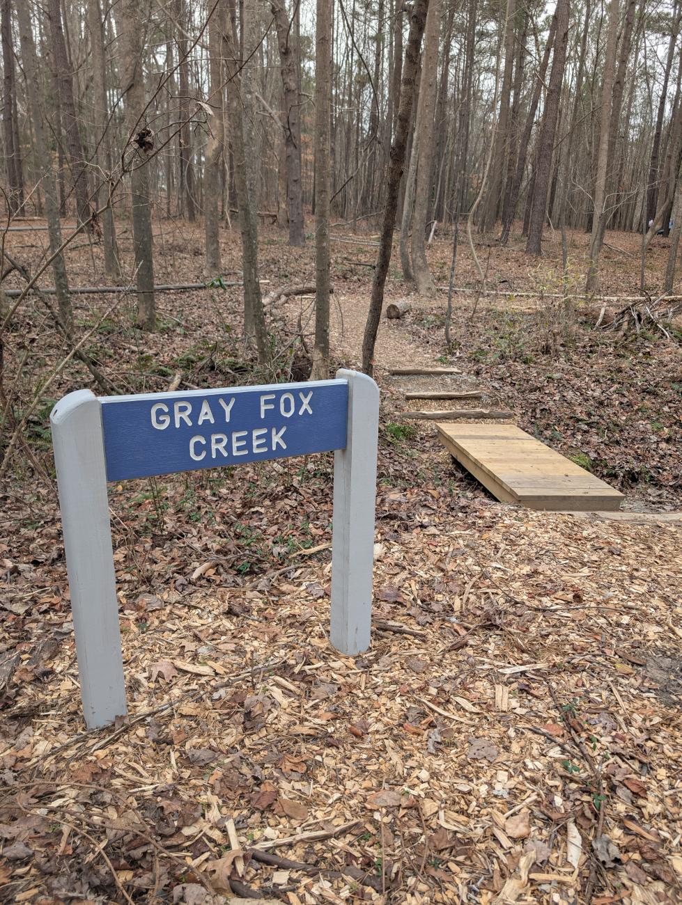 Photo of blue wooden sign with name Gray Fox Creek Trail next to wooden footbridge and path through woods