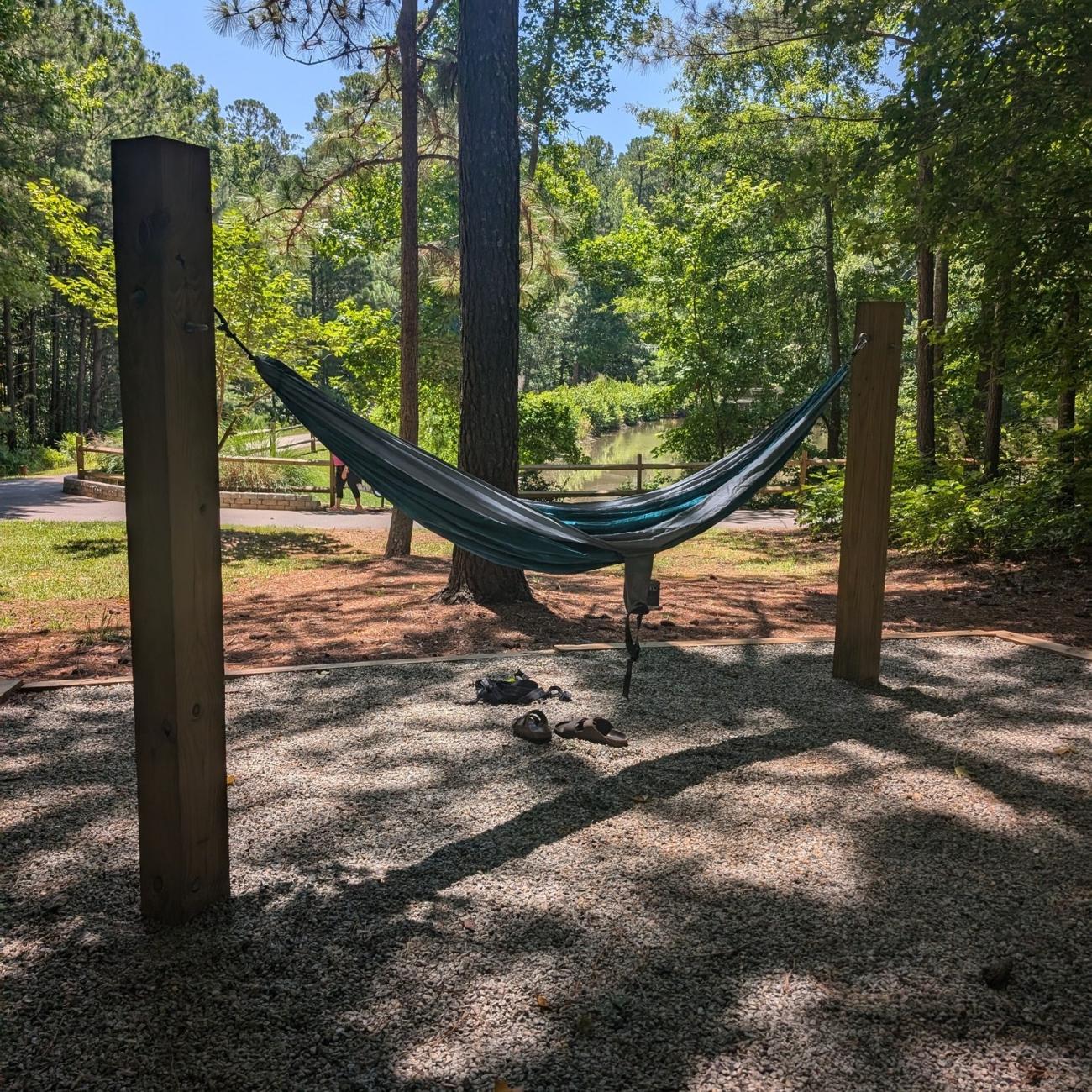 A hammock hangs from 2 upright wooden beams that are embedded in a gravel covered area