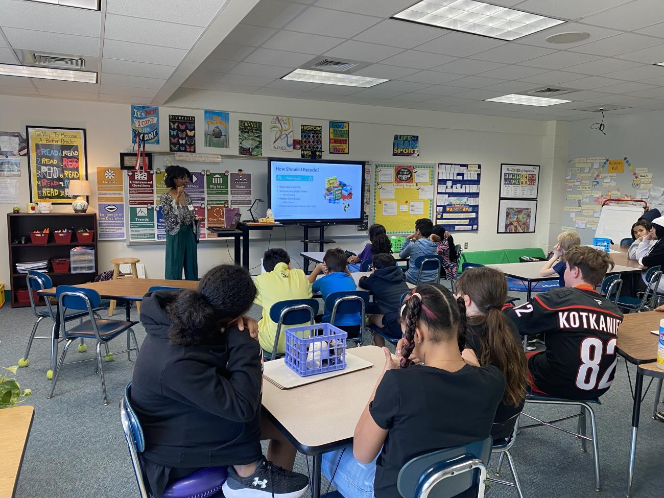 A woman stands next to a large computer screen in a classroom of children