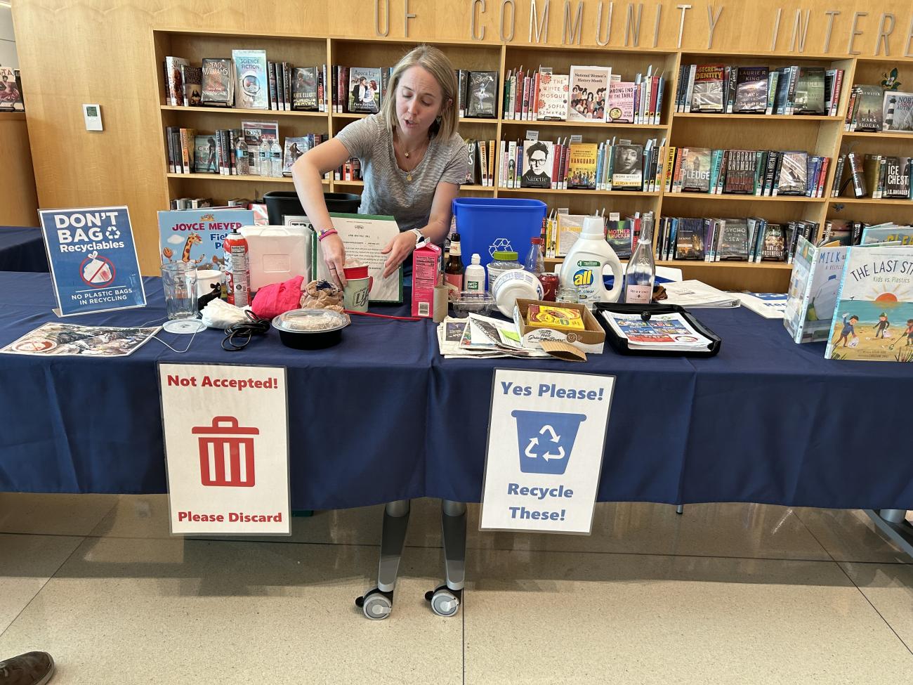 A woman sets up a table with examples of recyclable and nonrecyclable items. Signs on the table read “Not Accepted! Please Discard” on the left and “Yes Please! Recycle These!” on the right. The display includes bottles, cardboard and food containers.