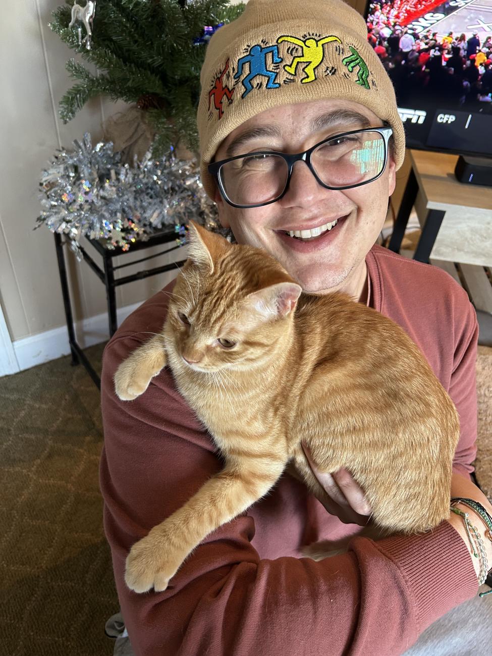 Smiling person wearing glasses, a rust-colored sweatshirt, and a light brown beanie with colorful dancing figures holds an orange tabby cat in their arms. A decorated Christmas tree and a TV showing a sports game are in the background.