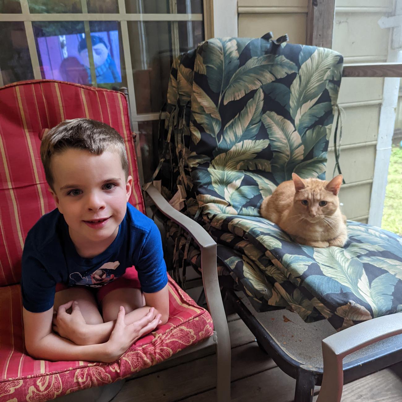 Sam, a young boy, sitting on a red chair next to Cheese, an orange tabby cat, who is lounging on a nearby chair – both on a screened porch.