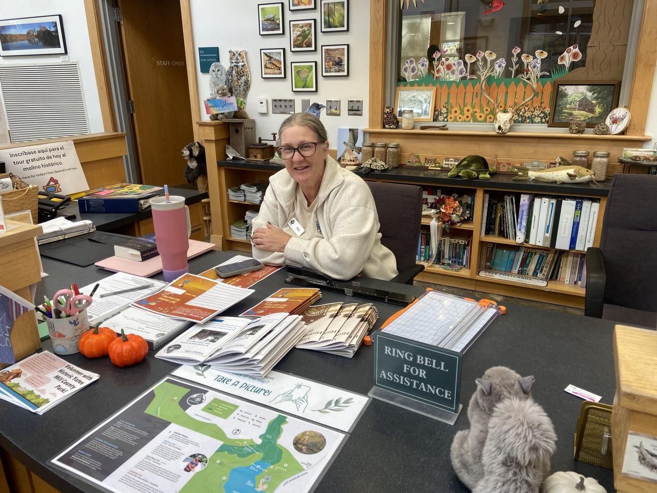A woman wearing glasses sits smiling with her arms crossed. She sits behind a large desk covered in brochures.