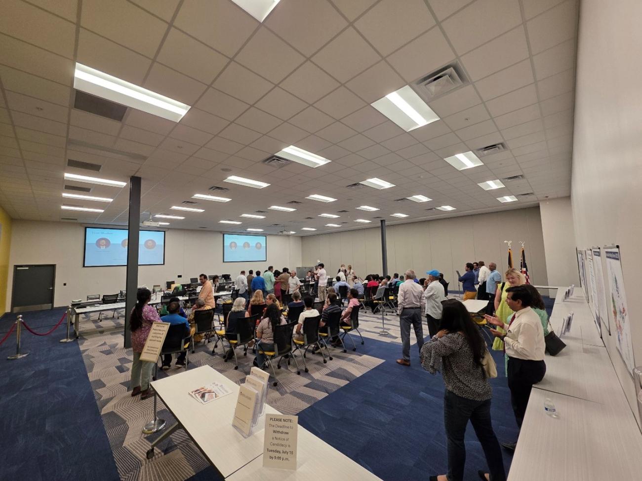 Group of people walking and sitting in a room with tables and signs. Images are projected on the walls.