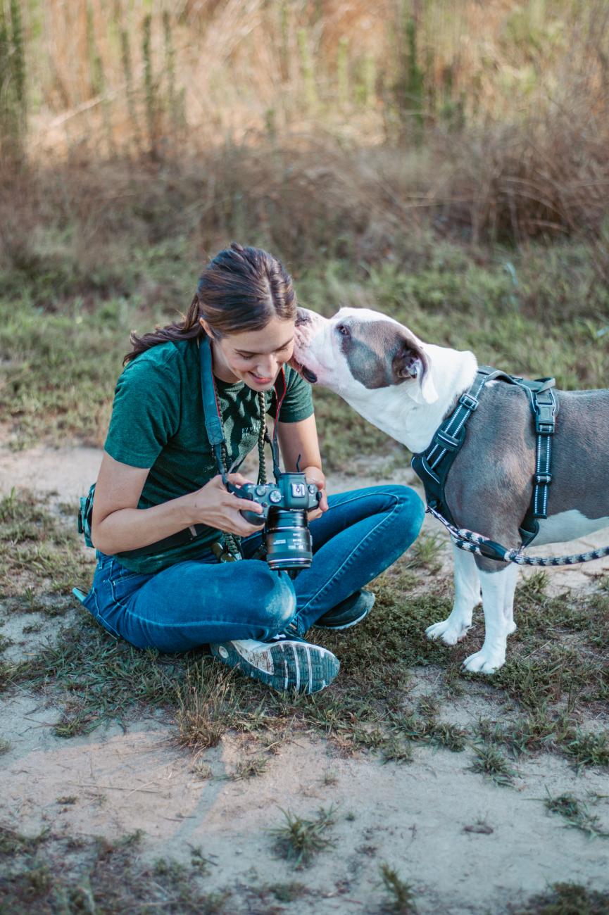 A woman sitting on the ground outdoors, smiling while holding a camera as a gray and white dog wearing a harness affectionately licks her face. The scene is set in a grassy, sunlit area.