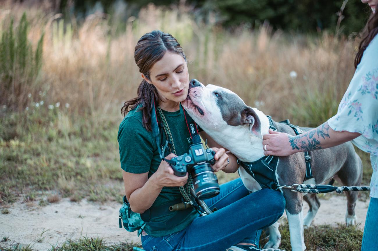 A woman sitting outdoors holds a camera while a large gray-and-white dog affectionately licks her face. Another person with a floral tattoo on their arm holds the dog’s leash. The scene takes place in a grassy field with warm, natural light.