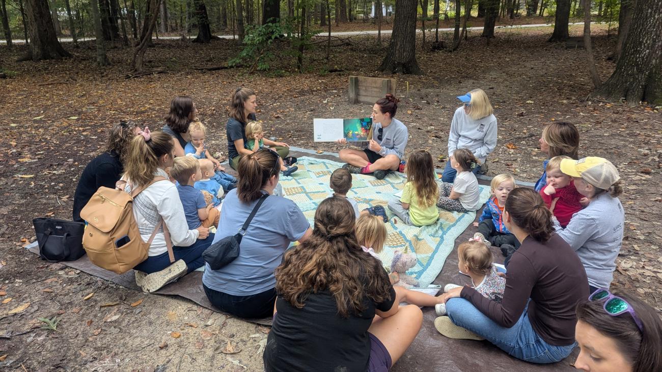 Group of children and caregivers gathered around staff member reading a book 