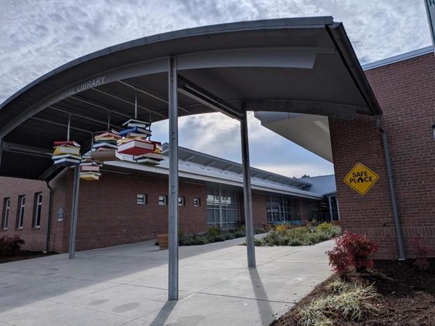 Entrance of a public library with a covered walkway and a hanging sculpture of colorful books. A yellow ‘Safe Place’ sign is mounted on the brick wall.
