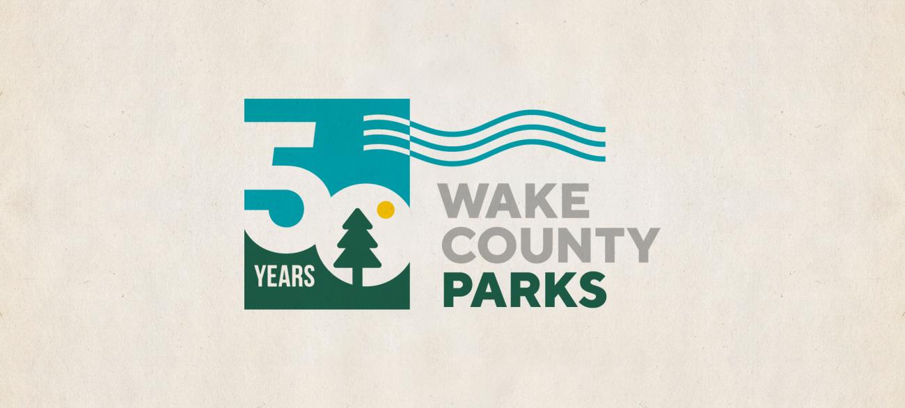 Commemorative logo celebrating 50 years of Wake County Parks, featuring a stylized number 50 with a tree, sun, water waves, and the words ‘Wake County Parks’ in green and gray on a light textured background.