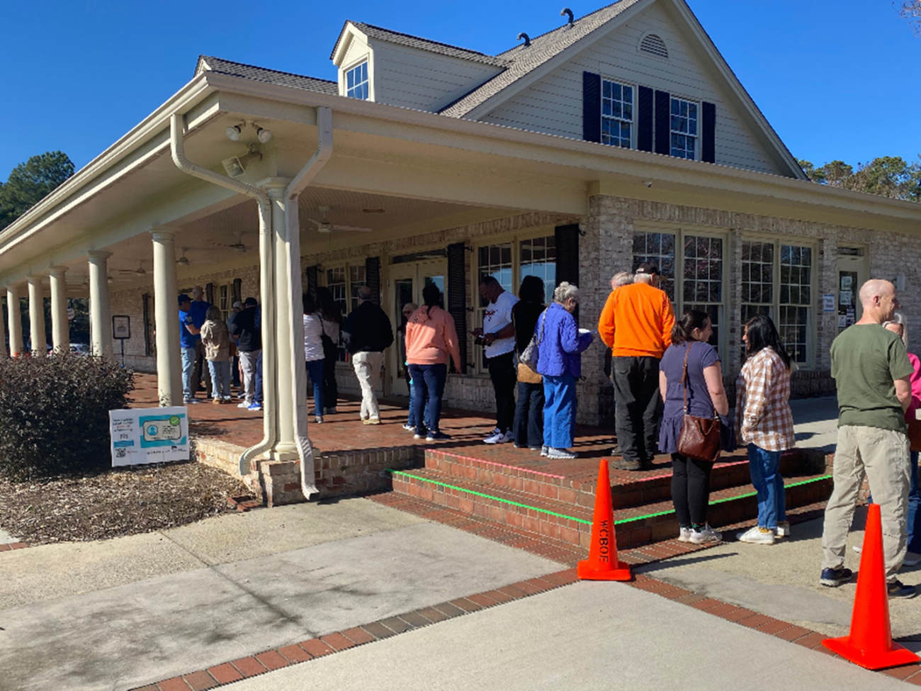 Voters stand in line outside an early voting location, waiting to enter a brick building with a covered porch on a clear day. Orange cones mark the walkway near the entrance.