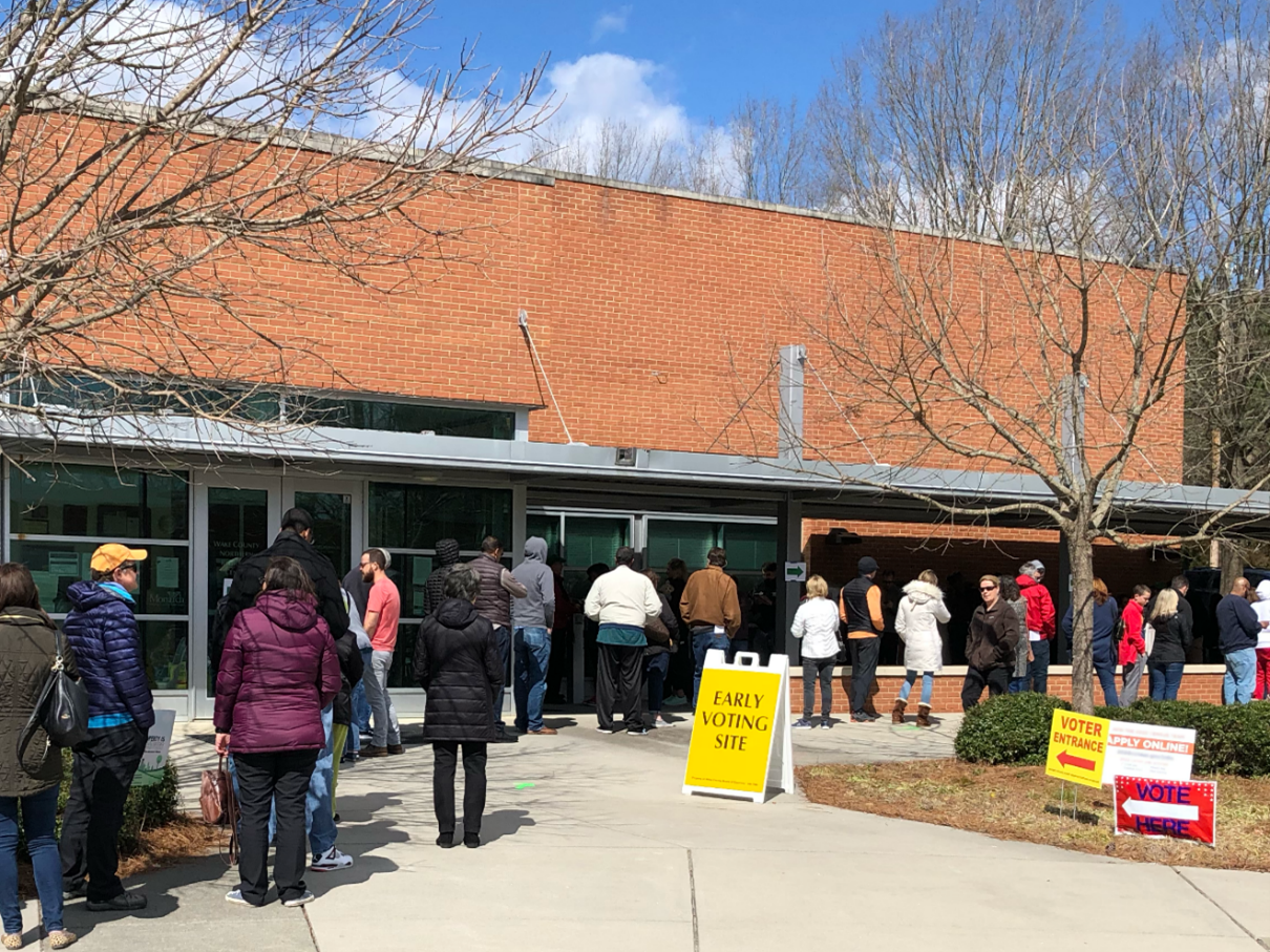 **Alt text:** People standing in line outside a brick building marked as an early voting site.