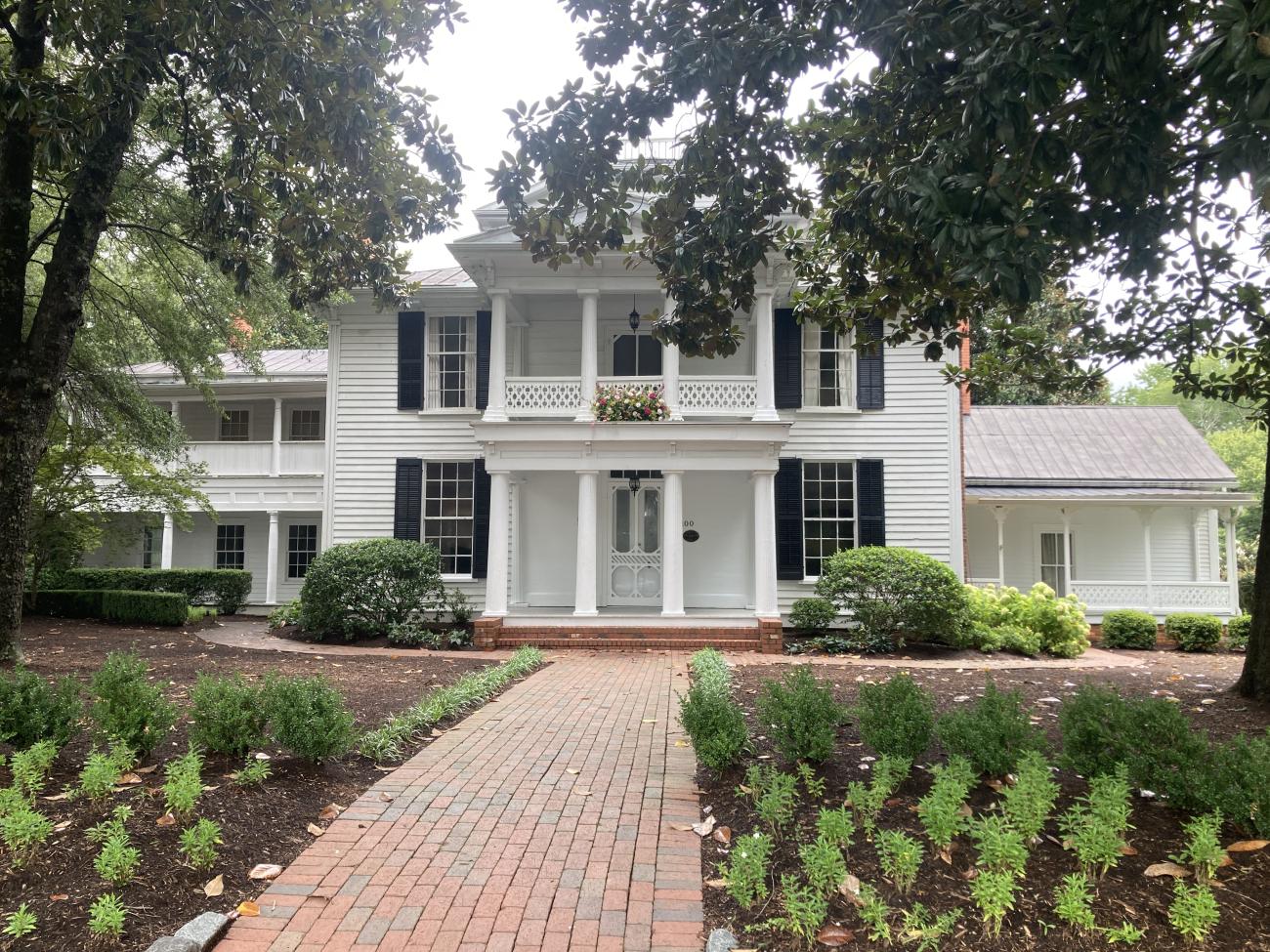 Large white historic home with black shutters, tall columns, and a brick walkway leading to the front entrance, surrounded by trees and landscaped gardens.