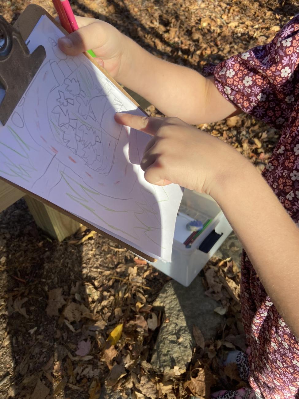 Close-up image of a child's hands holding a clipboard and pointing at a drawing. The drawing depicts a child's map of a trail surrounded by trees.