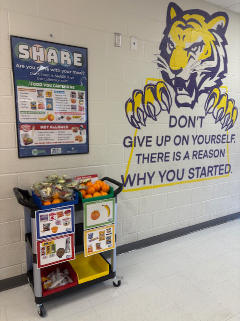 Project SHARE food cart with fruit and packaged snacks beneath a “Share” food donation poster, next to a wall mural of a tiger and the message “Don’t give up on yourself. There is a reason why you started.”