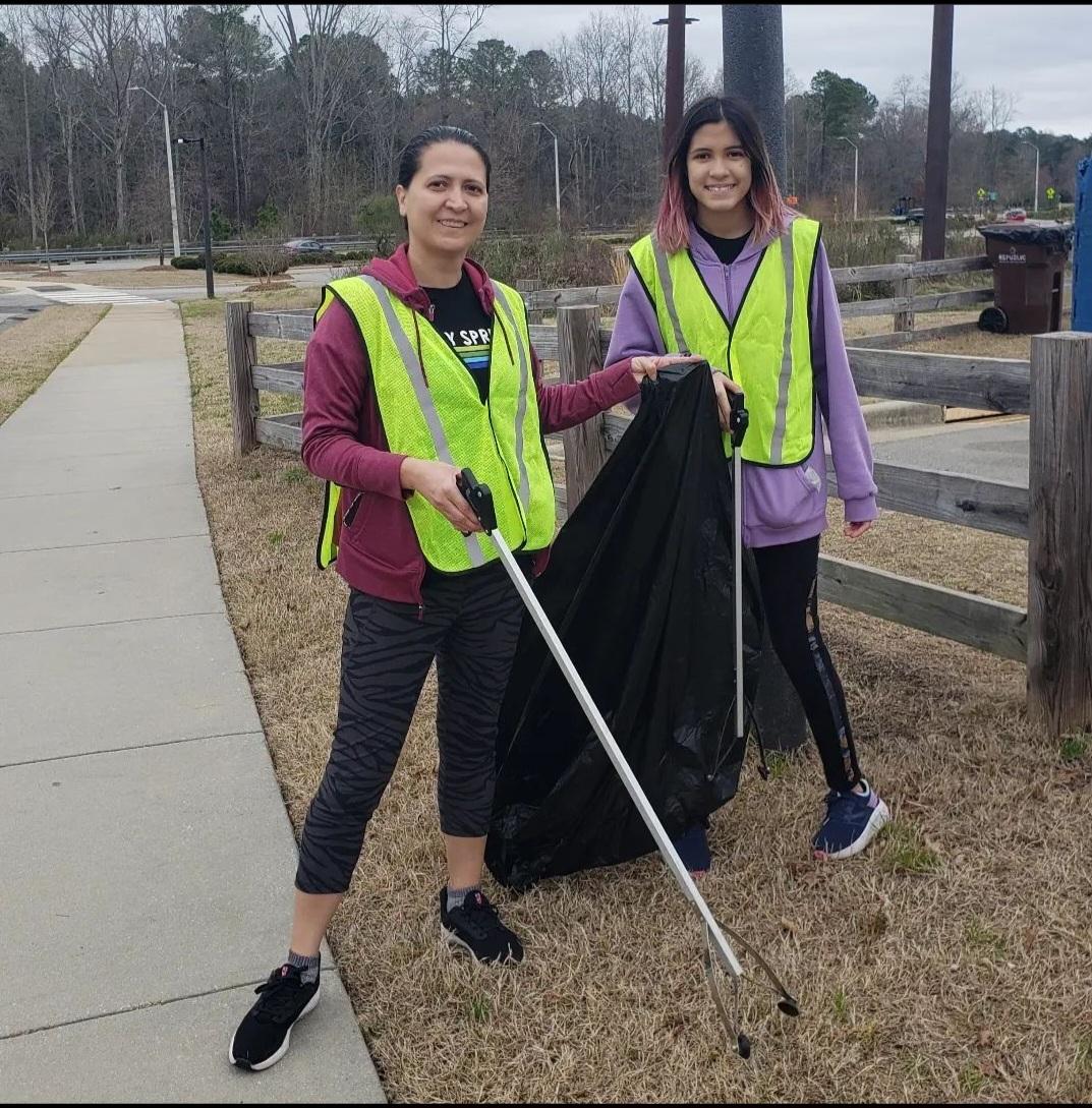 Two volunteers wearing safety vests collect roadside litter with grabbers and a trash bag along a sidewalk during a community cleanup.