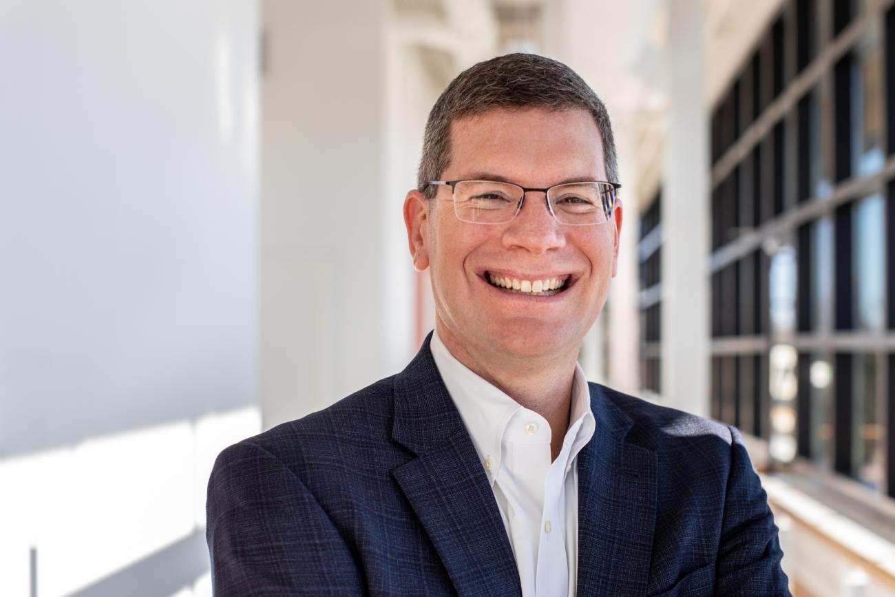 Man wearing glasses and a blazer smiles at the camera in a bright indoor hallway.