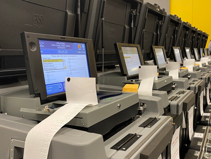 Row of voting machines with screens and printed ballots in a polling or election processing area.