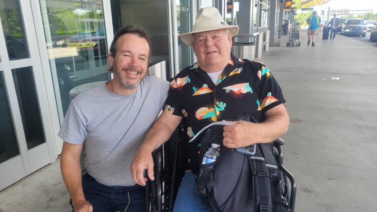 Two men smiling together outside a building, one seated in a wheelchair holding a bag.