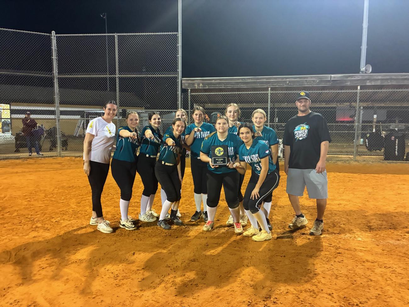 Youth softball team posing on a field at night with coaches, holding a trophy.