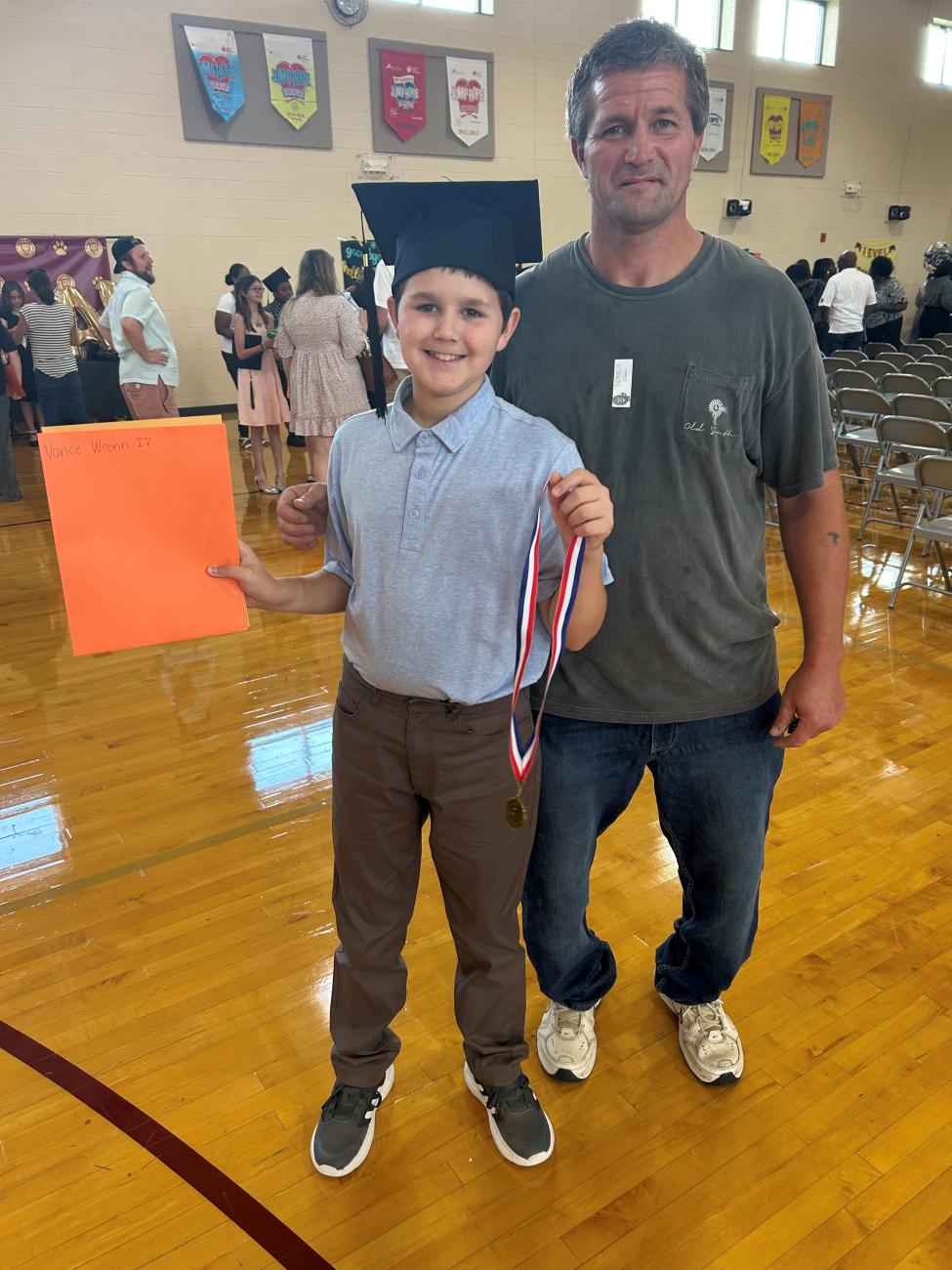 Boy in a graduation cap holding a certificate and medal, standing with an adult in a gym.