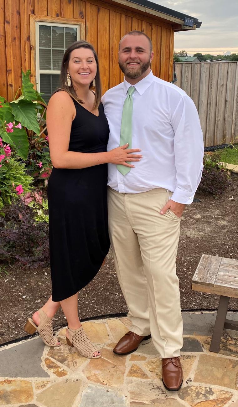 Couple dressed nicely posing together outdoors in front of a wooden building and garden.
