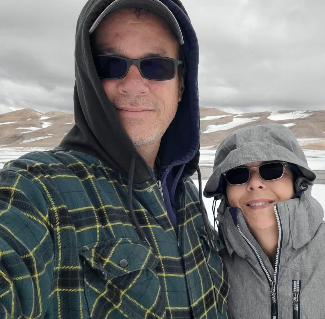 Two people in hooded jackets and sunglasses taking a selfie in a snowy landscape.