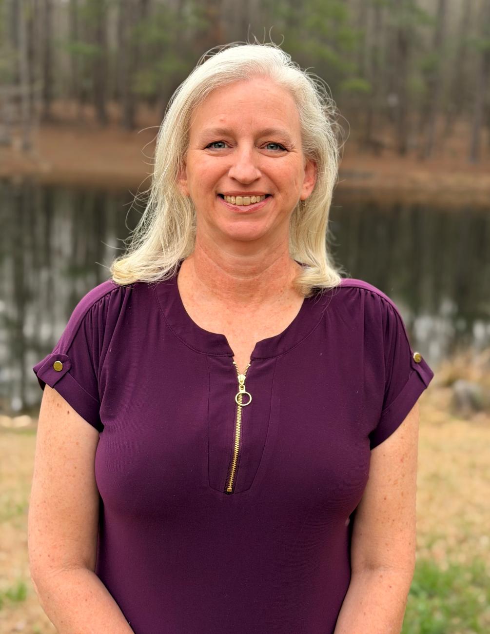 A smiling woman with light hair wearing a purple top, standing outdoors near a calm body of water with trees in the background.