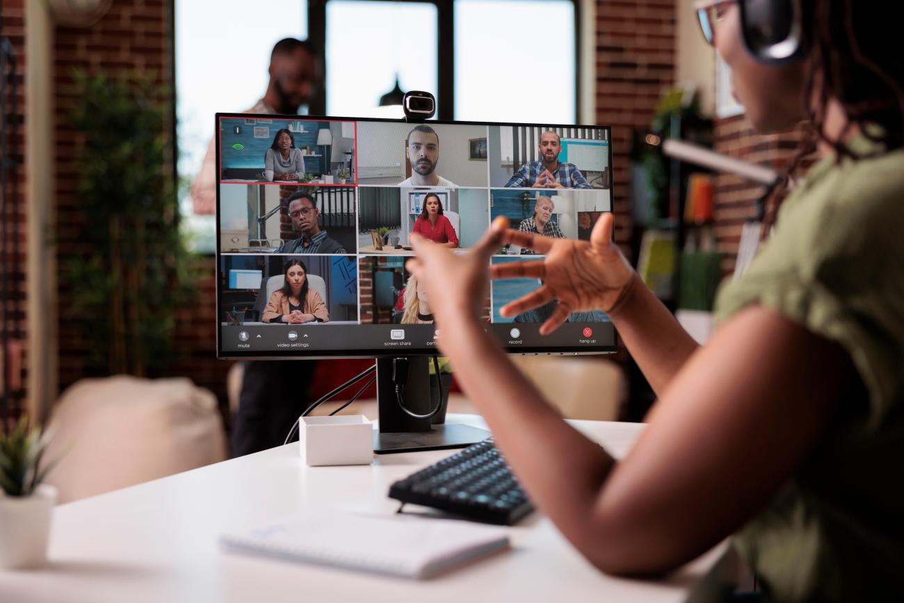Person wearing a headset participates in a video conference, speaking to multiple people shown on a computer screen.