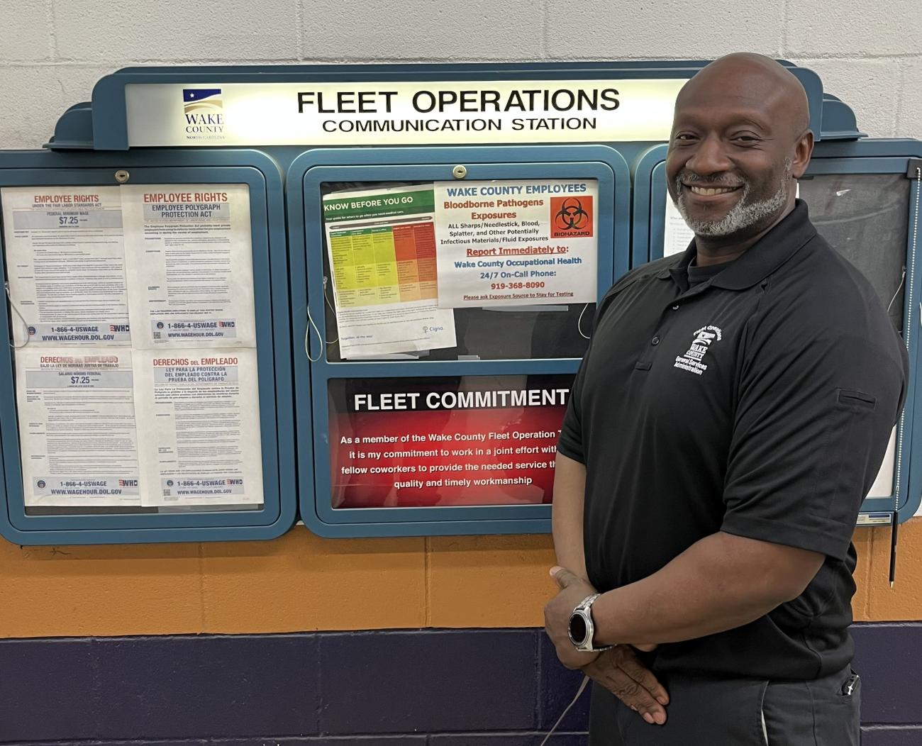 Man stands smiling beside a “Fleet Operations Communication Station” board displaying employee notices.
