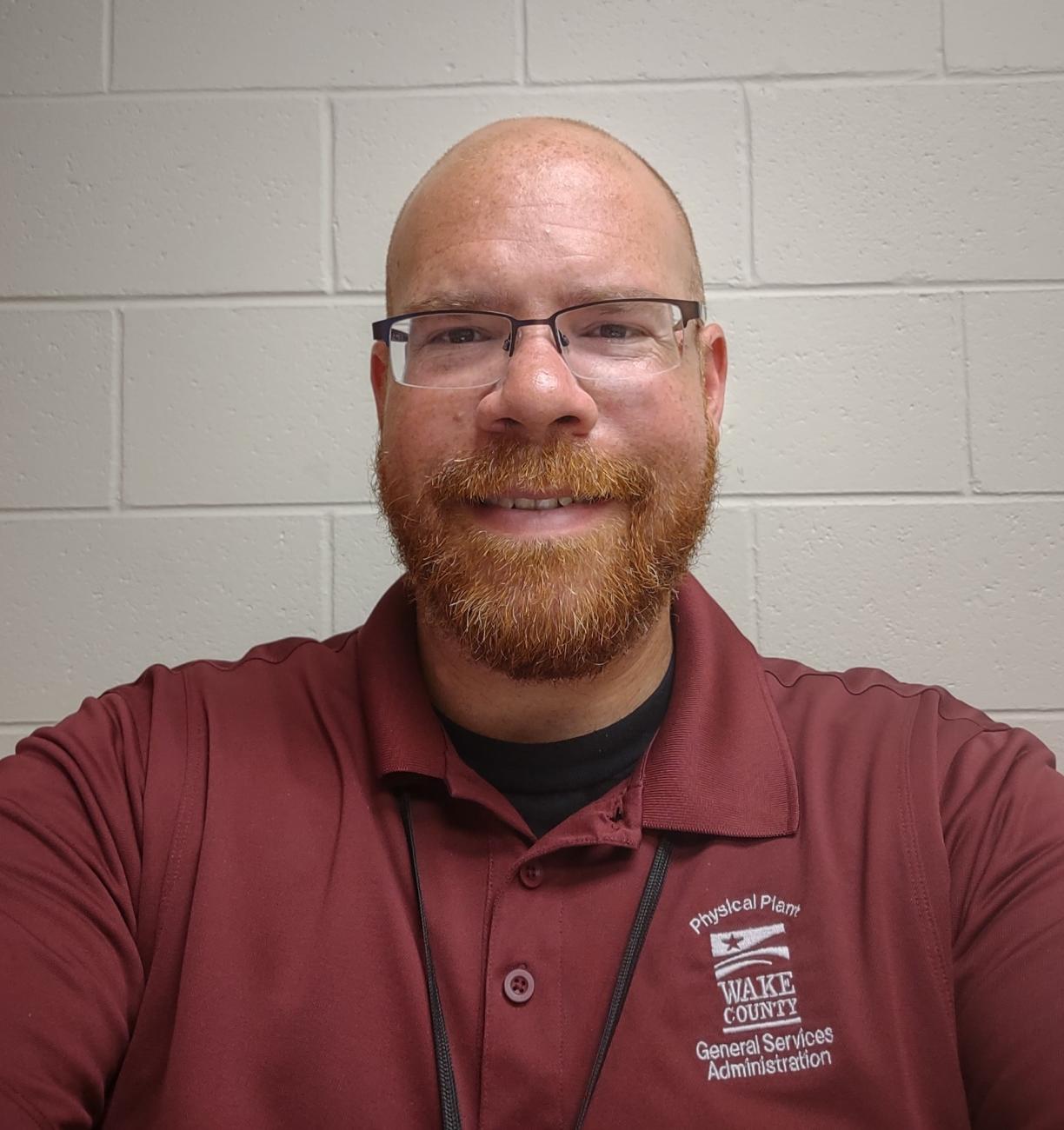 Man with glasses and a beard smiles at the camera against a plain wall, wearing a Wake County General Services shirt.