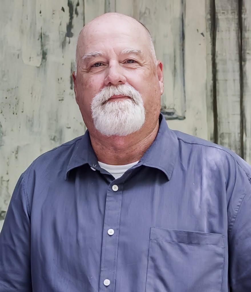 Older man with a bald head and white beard, wearing a blue button-up shirt, standing in front of a weathered wall.
