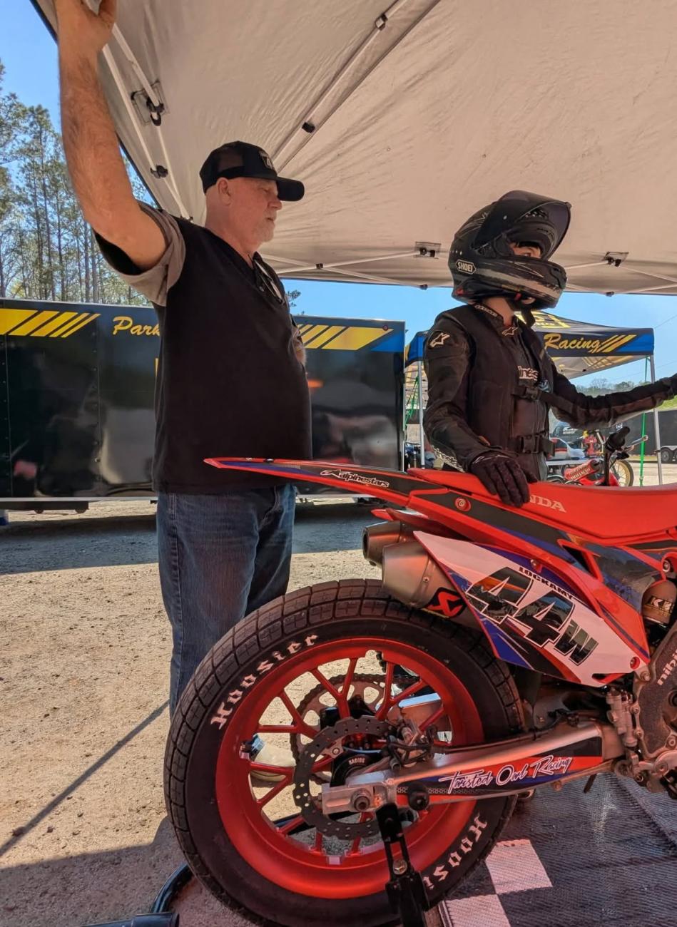Man standing under a canopy beside a child in full riding gear next to a red racing motorcycle.
