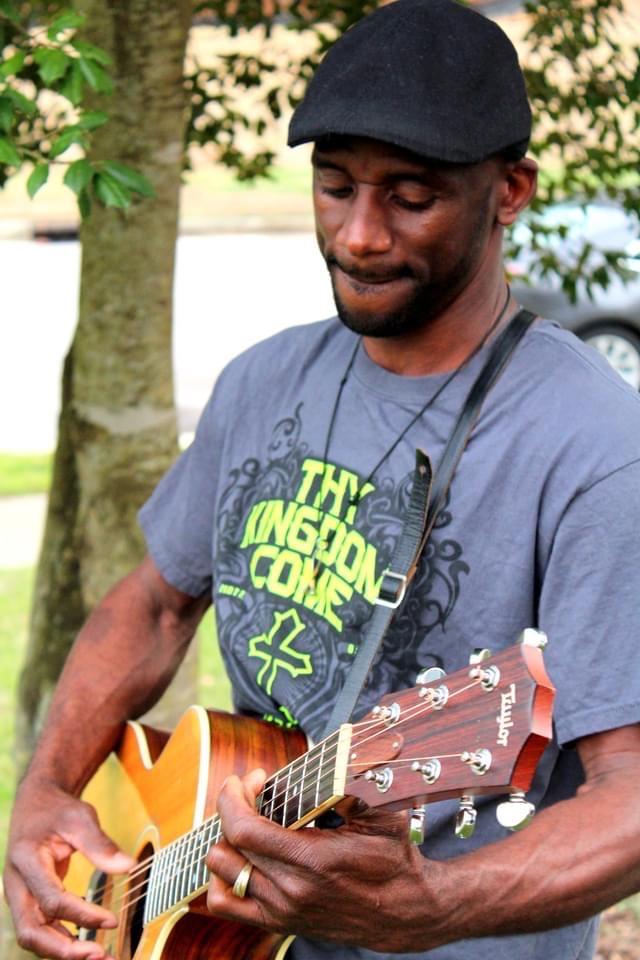Man wearing cap and a t-shirt with "Thy Kingdom Come" written on it plays a guitar in an outdoor setting.