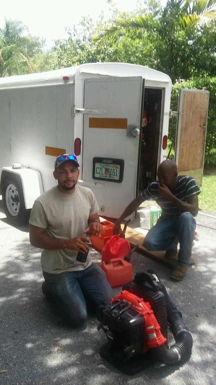 A man kneels on the pavement holding a small container, with a leaf blower in front of him. Nearby are red fuel containers.