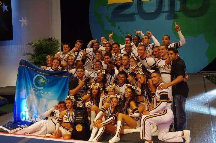 A large group of cheerleaders posing together on a stage, with medals around their necks, a banner that includes the words “Cheerleading Worlds,” and a trophy.