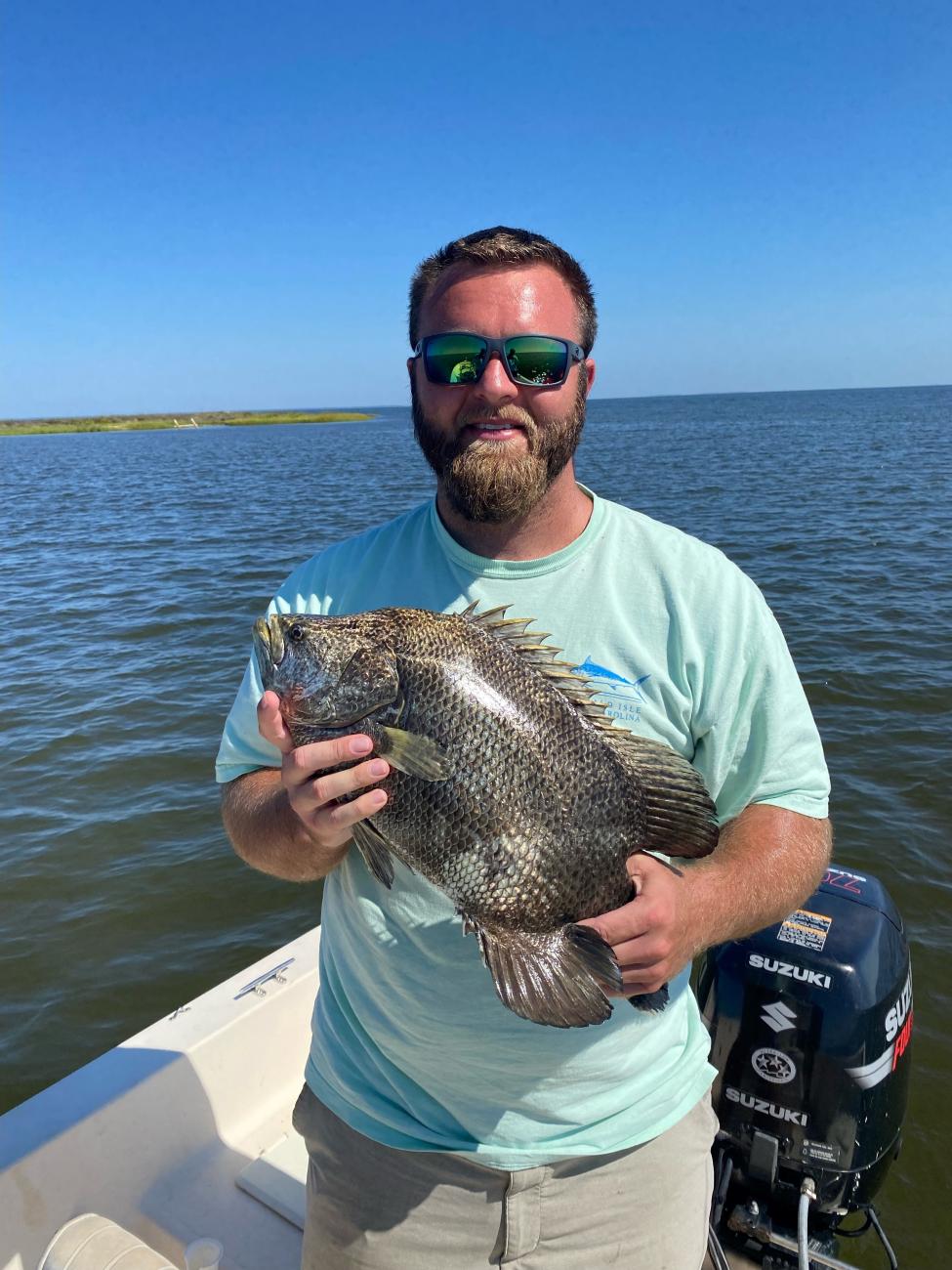 Man on a boat holding a large fish, with water and shoreline in the background.