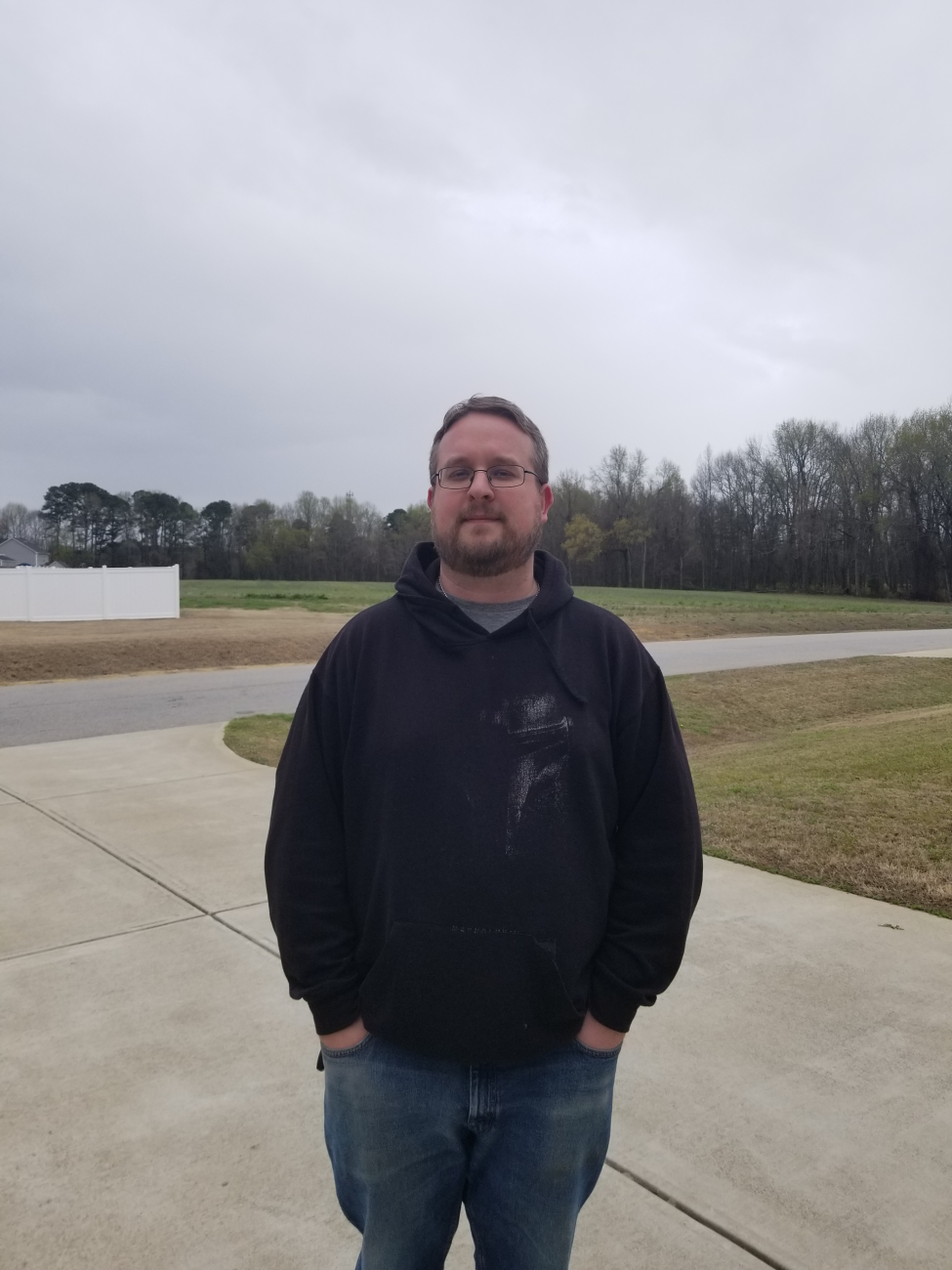 Man wearing glasses and a hoodie stands on a driveway in a residential area under a cloudy sky.