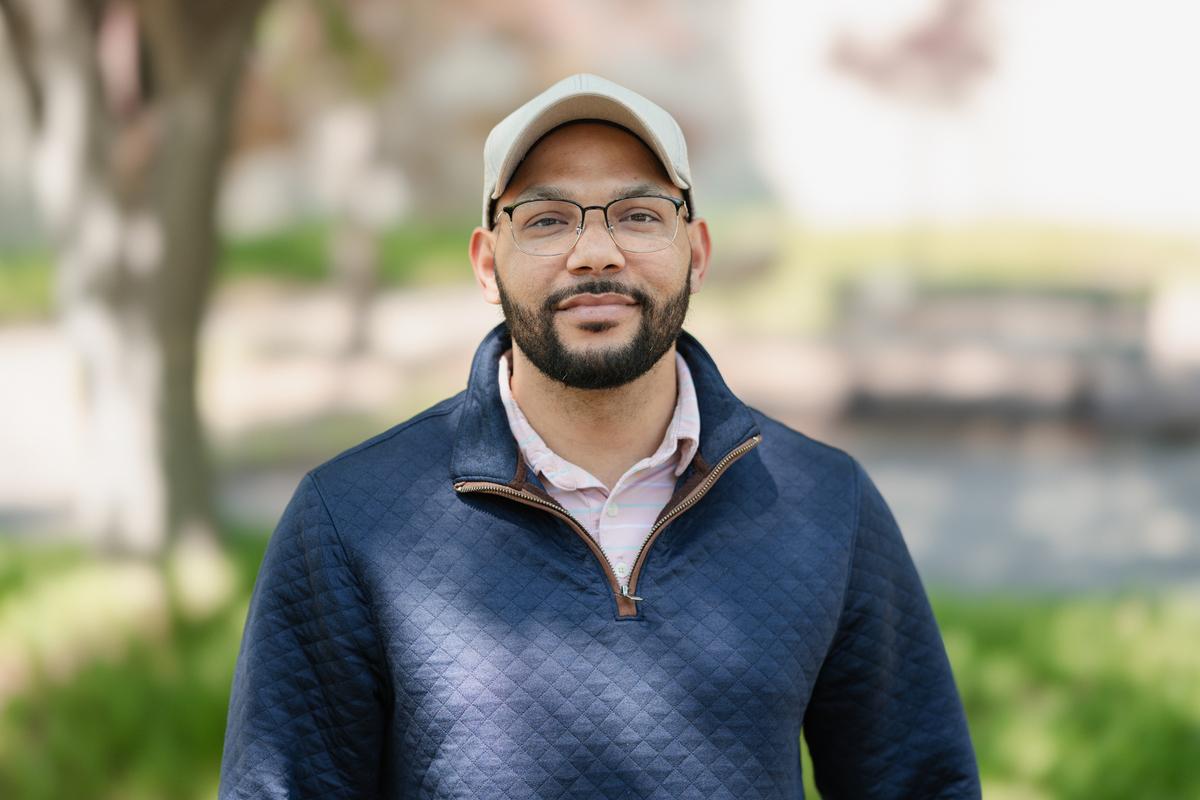 Smiling man with a cap, glasses and beard, wearing a thermal pullover, stands in a landscaped parking lot.