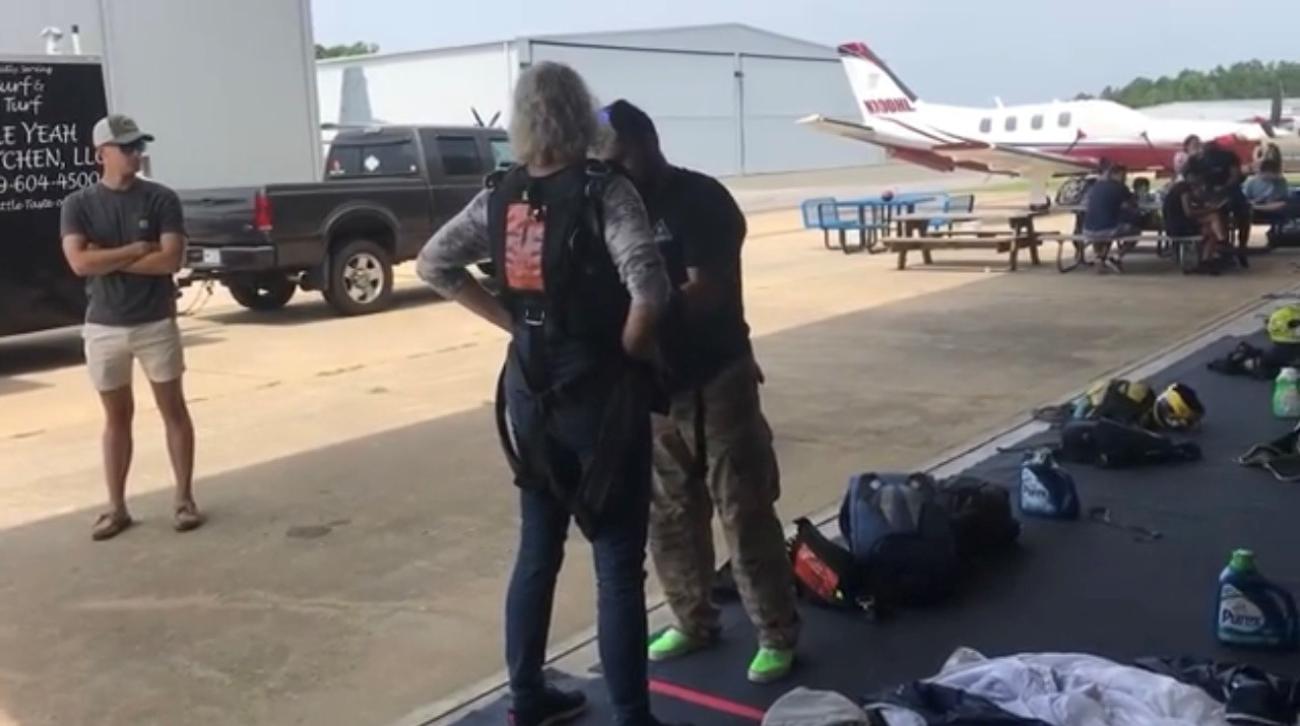 A small group of people at an airfield—one person is helping another into skydiving gear, while others stand nearby. A small airplane and equipment are visible in the background.