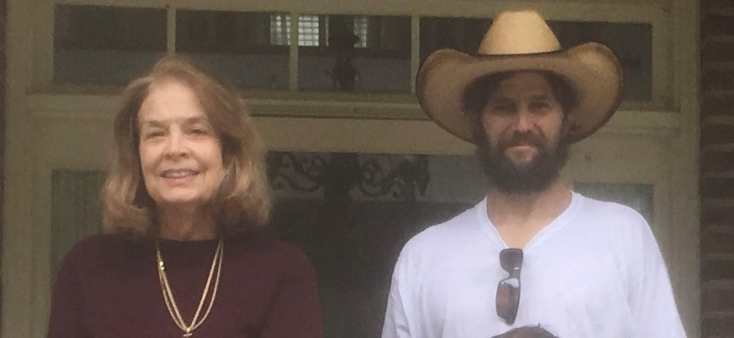 Two people standing together in front of a house, one wearing a cowboy hat.