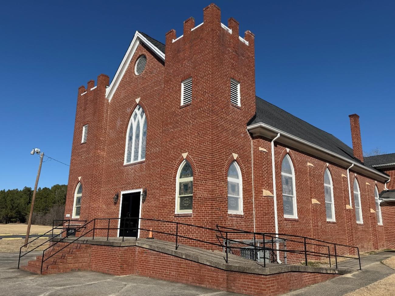Red brick church with arched windows and a ramped entrance under a clear blue sky.