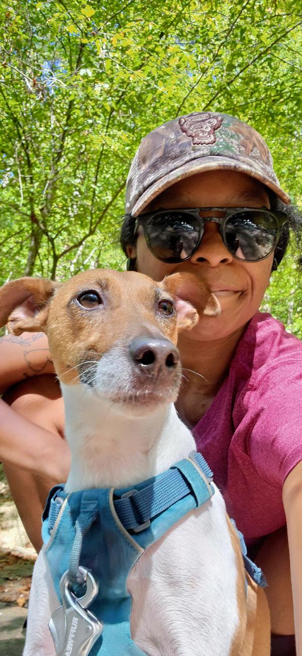Person wearing sunglasses and a cap poses with a small dog in a blue harness on a wooded trail, with green trees in the background.