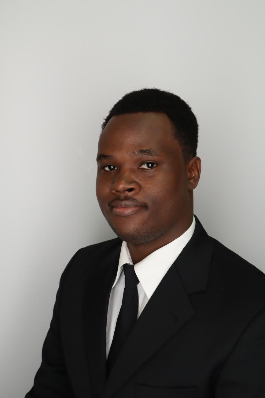 Young man in a black suit and tie poses for a professional headshot against a light gray background.