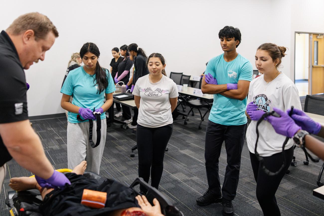 A group of young EMS interns watch as their EMS instructor demonstrates stop-the-bleed techniques on a dummy that is lying on a stretcher