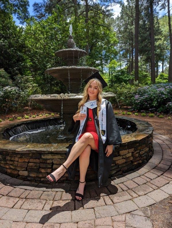 Graduate in cap and gown sits beside a stone fountain, smiling outdoors on a sunny day.