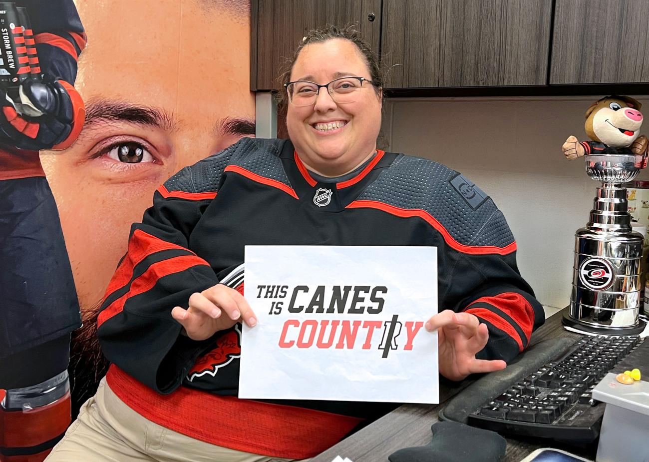 Smiling person in a Carolina Hurricanes jersey holds a “This Is Canes Country” sign while seated at an office desk with Hurricanes-themed decor.