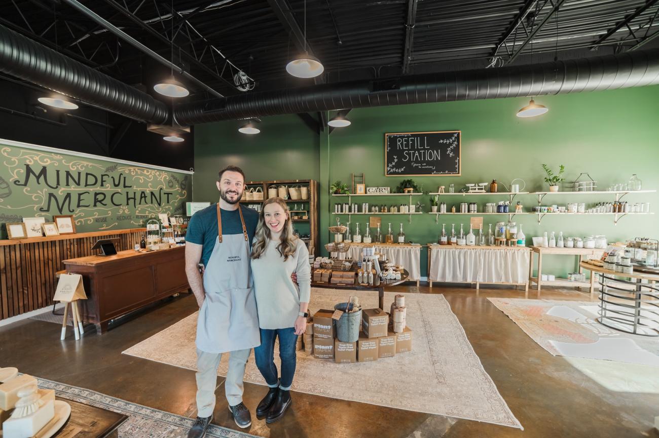 Two people stand smiling inside a refill and sustainable goods store, surrounded by shelves of reusable containers, household products, and eco-friendly merchandise.