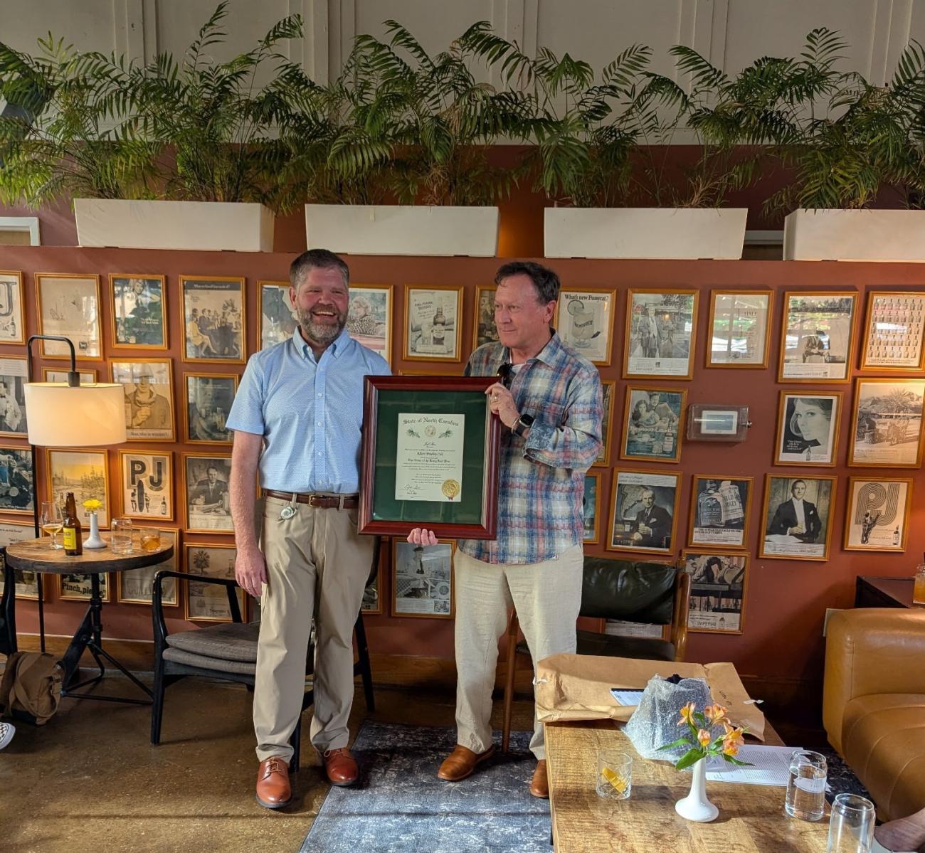 Two men stand together while one presents the other with a framed Order of the Long Leaf Pine certificate during an indoor recognition event.