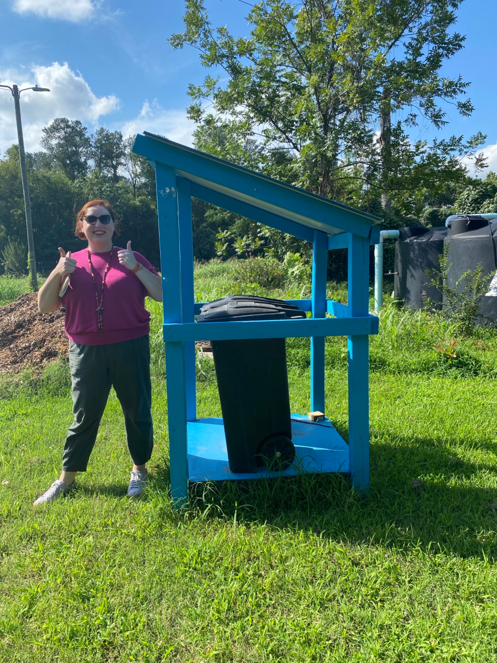 Smiling woman gives two thumbs up beside a bright blue shelter containing a trash cart in a grassy outdoor area.